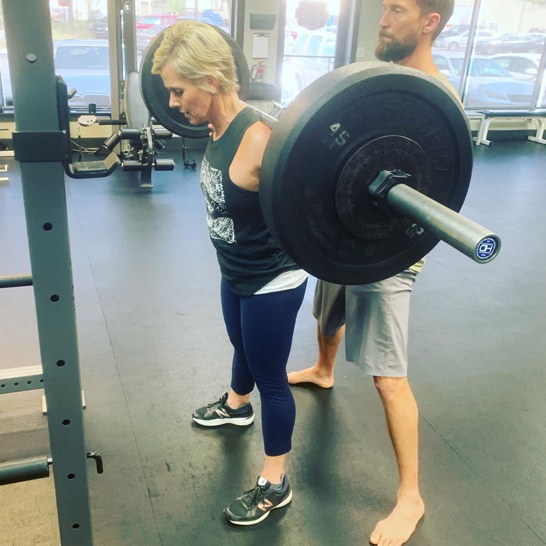 A coach stands behind a member as she sets up for a barbell back squat.