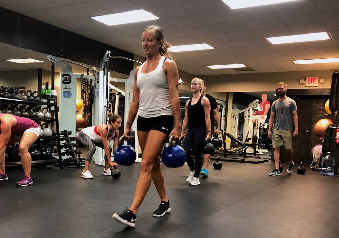 A member carries two kettlebells across the gym while others train behind her.