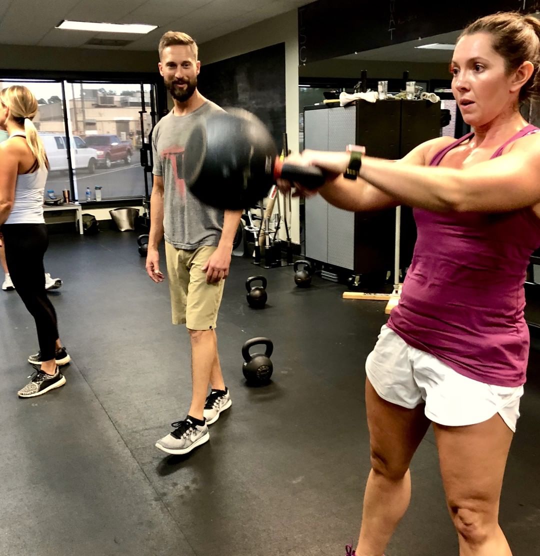 A coach watches a member swing a kettlebell during a group class.